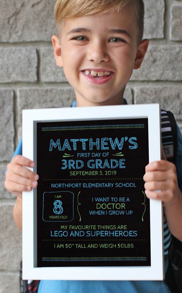 Boy holding framed first day of school sign with blue and green chalkboard design